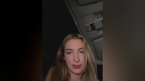 Media: A video of a young woman with long, blonde hair and light skin, wearing minimal makeup, captured in a dimly lit airplane cabin.