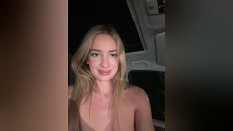 Media: Video of a fair-skinned, blonde woman with wavy hair, wearing a light-colored top, smiling in the dimly lit interior of a vehicle.