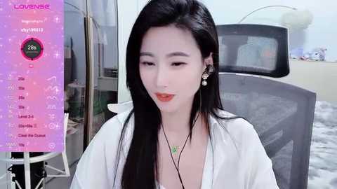 Media: A video of a young East Asian woman with long black hair, fair skin, and red lipstick, sitting in a chair, wearing a white top and pearl earrings, with a background featuring a car and a TV screen.