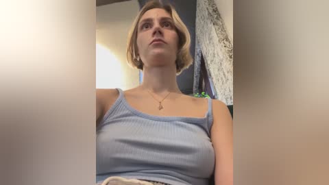 Media: Video of a fair-skinned woman with short blonde hair, wearing a light blue ribbed tank top, sitting in a room with a textured wall and hanging clothes.