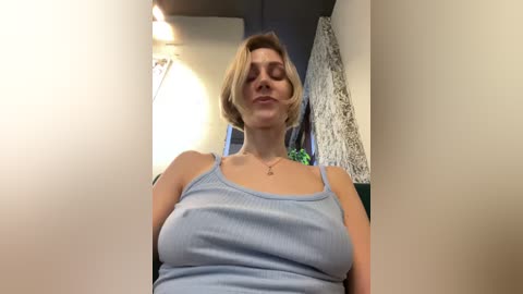 Media: Video of a fair-skinned, blonde-haired woman with a slim figure, wearing a light blue ribbed tank top, sitting indoors with a neutral expression, surrounded by a modern, slightly dimly lit room.