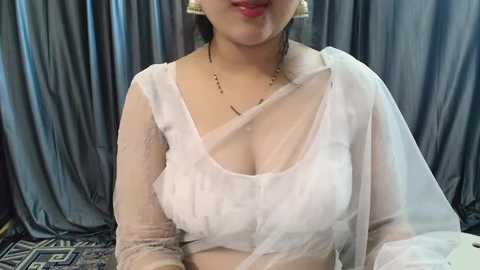 Media: Video of a South Asian woman with fair skin and medium-sized breasts wearing a sheer white blouse, draped with a white sari, against gray draped fabric.