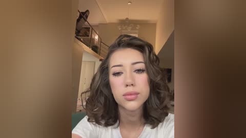 Media: Video of a young Asian woman with wavy, shoulder-length brown hair, light skin, and full lips. She wears a white t-shirt in a modern, warmly lit living room with a staircase and chandelier.