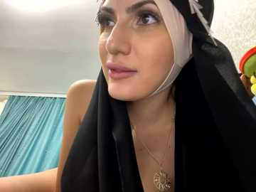 Media: A video of a young woman with fair skin and dark eyes, wearing a black hijab and gold necklace, standing in a room with teal curtains and a white wall.