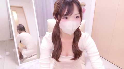 Media: Video of an East Asian woman with long brown hair in pigtails, wearing a white mask and white top, sitting at a white table with a mirror reflection showing her backside. The background features white walls and wooden furniture.