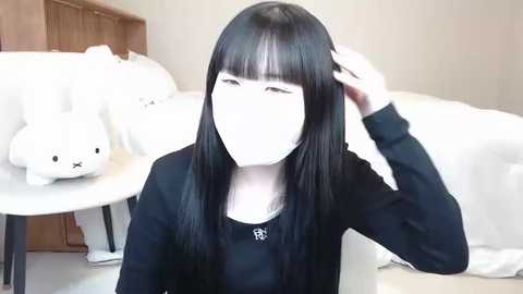 Media: A video of a young East Asian woman with long black hair and a black sweater, adjusting her hair in a bedroom with white bedding and a stuffed rabbit on a wooden table.