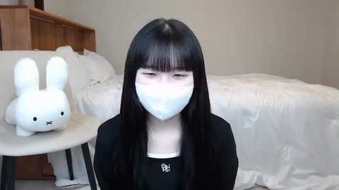 Media: Video of a young woman with long black hair, wearing a white mask, black sweater, and seated on a bed in a simple, modern bedroom with white sheets, a wooden dresser, and a stuffed white rabbit.