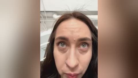 Media: Video of a young woman with fair skin, dark hair, and green eyes, taken from a low angle, in a bathroom with a white bathtub and white wall in the background.