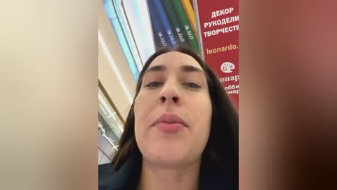 Media: Video of a woman with straight, shoulder-length brown hair, light skin, and closed eyes, standing in a library with bookshelves and a red sign in the background.