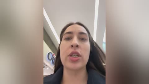 Media: A video of a young woman with long dark hair, fair skin, and light pink lipstick, wearing a black blazer, captured from a low angle with blurred objects in the foreground.