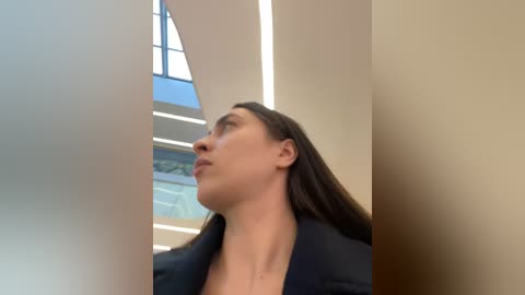 Media: Video of a Caucasian woman with long brown hair, wearing a black blazer, gazing upward through a modern, geometric skylight. The background features a minimalist interior with light, beige walls and a hint of a lush green landscape outside.