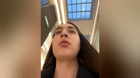 Media: Video of a young woman with long brown hair, looking upward, in a modern, brightly lit interior with a glass ceiling and decorative tiles.