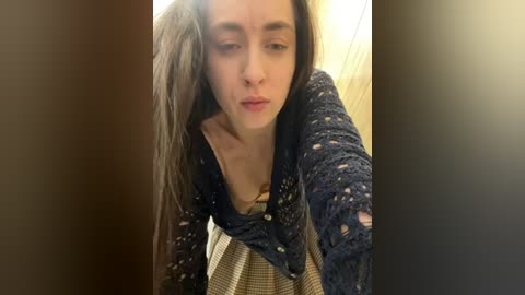 Media: A video of a young, fair-skinned woman with long, dark hair, wearing a blue, knitted cardigan over a striped top, leaning forward with a contemplative expression.