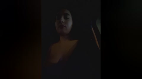 Media: A dimly lit video of a young woman with fair skin and short dark hair, wearing a black top, against a dark background.