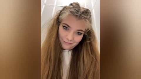 Media: Video of a young woman with long, straight, light brown hair, styled in a messy bun, smiling. She wears a white top and stands in a well-lit room with a white tiled wall in the background.