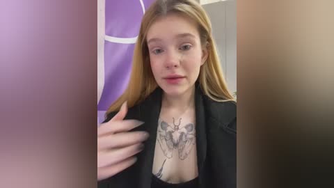 Media: Video of a young woman with fair skin and long blonde hair, wearing a black blazer over a black top, featuring a large butterfly tattoo on her chest. Background includes a purple wall with white circular designs.