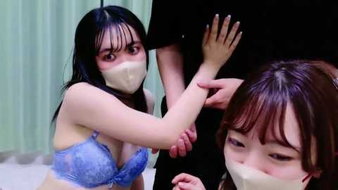 Media: Video of two Asian women in light blue lace lingerie and face masks, one restrained by another's arm, set in a dimly lit room with green curtains.