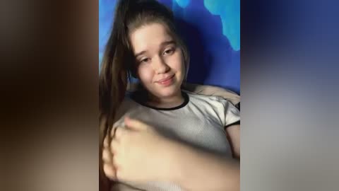 Media: Video of a young girl with long brown hair, fair skin, wearing a white T-shirt, lying on a blue background, smiling, arms around her torso.