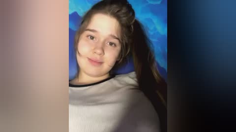 Media: Video of a young Caucasian girl with light skin, brown hair, and freckles, wearing a white t-shirt, against a blue and purple background. She has a slight smile and is leaning against a pillow.