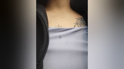 Media: Video of a woman's upper chest, showing a black floral tattoo and a light blue off-shoulder top, partially covered by a black leather jacket.