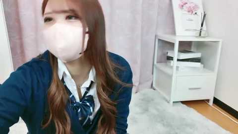 Media: Video of an East Asian woman in a navy school uniform, wearing a face mask, with long wavy brown hair. Background features a pink curtain, white shelving unit with books, and a flower painting.