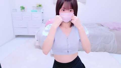 Media: Video of an East Asian woman with shoulder-length brown hair, wearing a light pink mask and a light gray cardigan, standing in a minimalist, white-walled bedroom with a neatly made bed and white dresser.