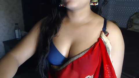 Media: Video of a South Asian woman with medium brown skin, wearing a red sari with blue bra peeking through, earrings, and dark hair. Background shows a dimly lit room with a TV and furniture.