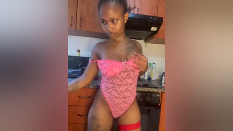 Media: Video of a dark-skinned woman with a slender, athletic build in a kitchen. She wears a pink lace bodysuit, red thigh-high stockings, and a black tattoo on her left thigh. The kitchen background includes wooden cabinets and a stove.