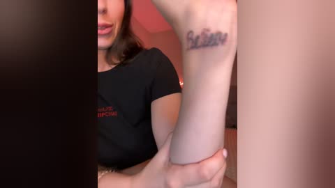 Media: Video of a woman with fair skin and brown hair, wearing a black shirt, holding her own arm. Her tattoo reads \"Be Free\" on the inner forearm.