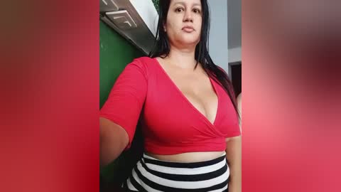 Media: A video of a plus-sized woman with long black hair, wearing a red crop top and black and white striped pants, standing in a kitchen with green and white walls.