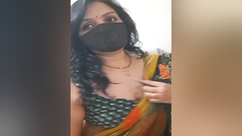 Media: A video of a woman with medium-dark skin, wearing a black face mask, black top, and orange sari, her wavy black hair partially visible.