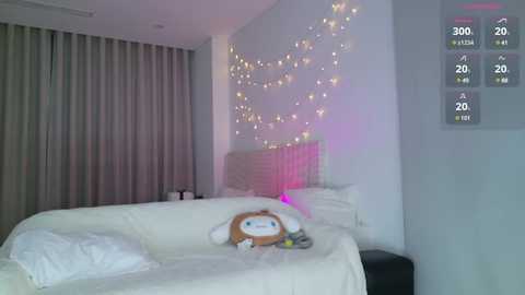 Media: Video of a minimalist, softly lit bedroom with a plush white bed adorned with a plush toy, a white nightstand, and a wall adorned with fairy lights.