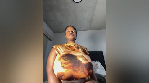 Media: A video of a Black woman with medium skin tone, short hair, and a medium build, wearing a tan t-shirt with a Renaissance painting of a woman. The background shows a dimly lit room with a bed and a plain ceiling.