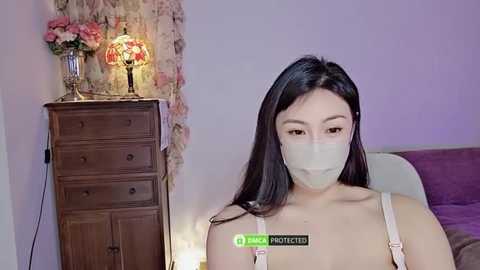 Media: Video of an Asian woman with long black hair, wearing a face mask, white tank top, and seated in a bedroom with a wooden dresser, floral wallpaper, and purple bedding.