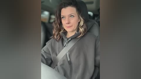 Media: Video of a middle-aged woman with shoulder-length wavy hair, wearing a grey hoodie, sitting in a car with a grey seatbelt, looking out the window.