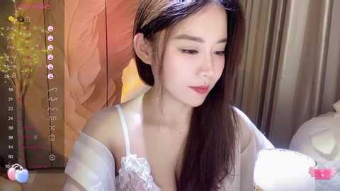 Media: A video of a young Asian woman with long, straight dark hair, wearing a white lace bra and a sheer white robe, seated in a softly lit room with wooden paneling and a floral arrangement.