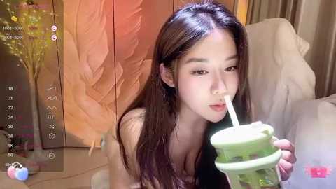 Media: Video of an Asian woman with long black hair, drinking from a green straw, wearing a black top, in a warm-toned, indoor setting with a tree and calendar visible.