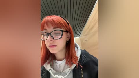 Media: Video of a young woman with straight red hair, glasses, black headband, black jacket, white hoodie, and gray shirt, standing indoors with beige walls and a black ceiling.