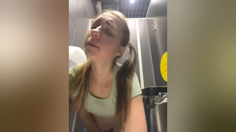 Media: Video of a young woman with long brown hair in pigtails, wearing a light green crop top, looking up with a neutral expression in a public restroom with metallic fixtures and a yellow circular sticker on the wall.