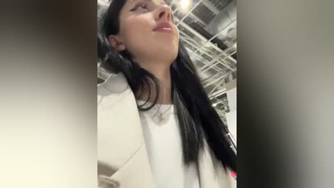 Media: Video of a young woman with long black hair, fair skin, and a white coat, looking up thoughtfully in a modern, industrial setting with exposed pipes and beams.