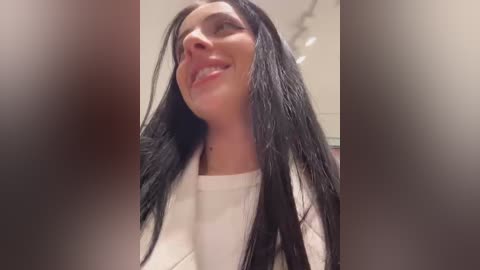 Media: Video of a smiling, light-skinned woman with long black hair, wearing a white top, in a blurry indoor setting with neutral colors and minimal visible details.