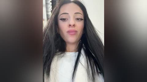 Media: Video of a young woman with long, straight black hair, wearing a white top, and a slight pout. Background is blurred, suggesting a hair salon setting.