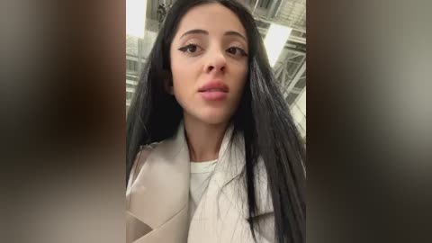 Media: Video of a young woman with long, straight black hair, wearing a white coat, standing indoors with industrial lighting.