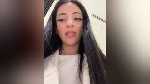 Media: A video of a woman with long, straight black hair, light skin, and full lips, wearing a white top, standing in a bathroom with white walls and a towel rack.