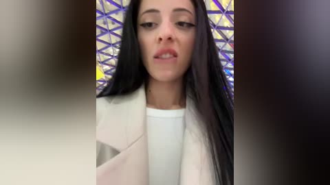 Media: Video of a woman with long black hair, wearing a white jacket, standing in front of a colorful geometric-patterned wall. She has a neutral expression and is indoors.