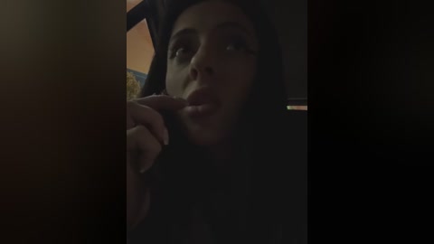 Media: A dimly lit video of a young woman with light skin, long dark hair, and full lips, seductively licking her finger. She wears a dark shirt, partially obscured by shadows. The background is indistinct, suggesting a private setting.