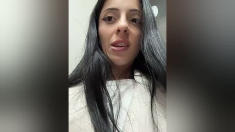 Media: A close-up video of a young woman with long, straight black hair and light skin, wearing a white shirt. She has a slight smile, and the background is blurred.