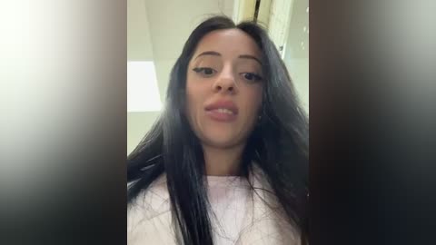 Media: Video of a young woman with long black hair, light skin, and full lips, wearing a white top, taken from a close-up angle indoors.