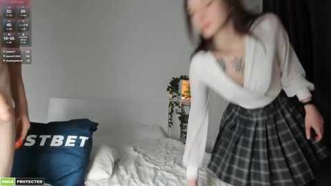 Media: Video of a woman in a white blouse and plaid skirt bending over a bed with a blue pillow labeled \"BET.\" Background features a white wall and a small table with a lamp.