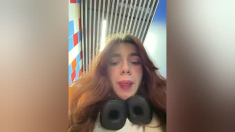 Media: Video of a woman with long, wavy auburn hair, wearing a red lipstick, lying on a bed with black headphones on her head, background features a colorful, striped wall.